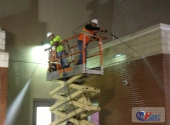 Two workers in hard hats and safety vests perform commercial pressure washing on a brick building using a scissor lift.